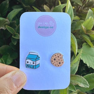 May include: A pair of stud earrings featuring a blue and white milk carton with a smiling face and a tan chocolate chip cookie with a bite taken out of it, both displayed on a white card with the text you can fly design co.