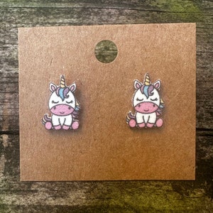 May include: A pair of unicorn stud earrings. The unicorns are white with pink, blue, and yellow details. They are sitting down with their eyes closed.