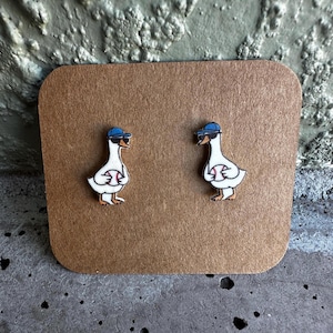 May include: A pair of stud earrings featuring a white goose wearing a blue baseball cap and black sunglasses while holding a baseball. The geese are illustrated with orange beaks and feet, presented on a rectangular brown cardboard display card.