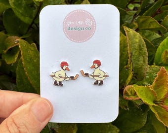 May include: A pair of stud earrings featuring a cartoon duck wearing a red hockey helmet and skates, holding a hockey stick and puck. The earrings are displayed on a white card with the text you can fly design co printed in a pink circle.