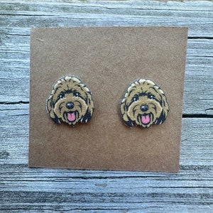 May include: A pair of brown and black fabric earrings featuring a doodle dog design. The dog has a pink tongue sticking out.