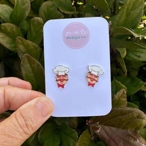 May include: A pair of stud earrings featuring the Swedish Chef character with a white toque, brown mustache, and red bowtie. The earrings are attached to a white display card with the text you can fly design co printed in a pink circle.