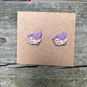 May include: A pair of purple and white cartoon dwarf earrings. The dwarf is wearing a purple hat and has a white beard.
