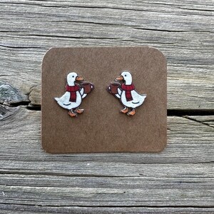 May include: A pair of duck earrings. Each earring features a white duck wearing a red scarf and holding a brown mug. The earrings are on a brown card, set against a wooden background.