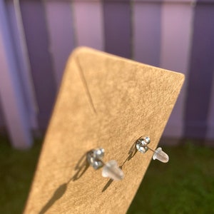 May include: A pair of silver stud earrings with clear plastic backs. The earrings are displayed on a brown card.