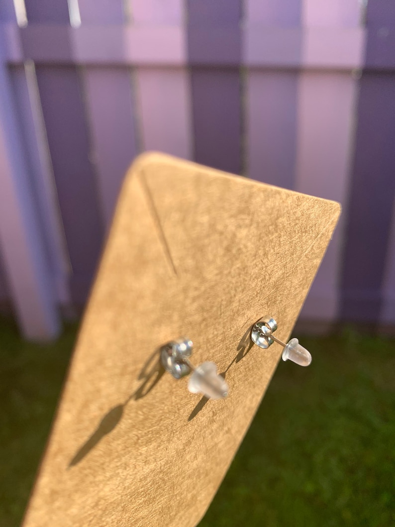 May include: A pair of silver stud earrings with clear plastic backs. The earrings are displayed on a brown card.