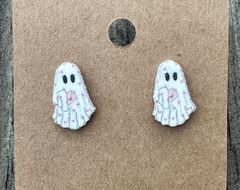 May include: A pair of stud earrings featuring white ghost shapes with small pink heart patterns. Each ghost figure has two black oval eyes and is holding a small pink envelope. The earrings are attached to a rectangular brown display card.
