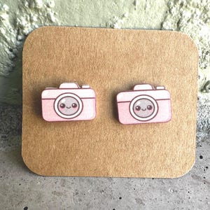 May include: A pair of stud earrings shaped like small pink cameras. Each camera features a central lens with a smiling face, including two black eyes and a small curved mouth. The earrings are displayed on a square piece of brown cardstock.