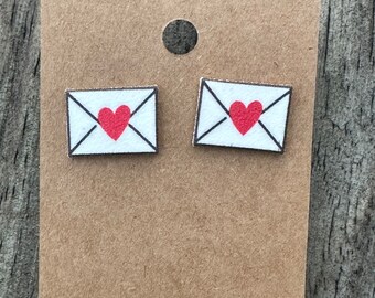 May include: A pair of stud earrings featuring a white envelope design with a red heart in the center. The earrings are displayed on a brown card, perfect for gifting or personal wear. The earrings are small and delicate.