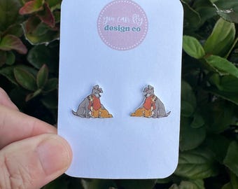 May include: A pair of stud earrings featuring a graphic of two dogs sitting together, displayed on a white card with the text you can fly design co in a pink circular logo. The dogs are brown and gray with detailed outlines.