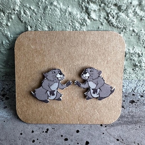 May include: A pair of gray groundhog earrings with white accents, set on a brown card. The groundhogs are depicted in a cartoon style, with their arms outstretched as if greeting each other. The background is a textured, light green surface.