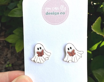 May include: A pair of white ghost stud earrings featuring a black outline and a brown book held by each ghost. The earrings are attached to a white display card with the text you can fly design co printed in a circular pink logo at the top.