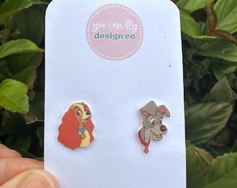 May include: A pair of stud earrings featuring the characters Lady and Tramp on a white display card with the text you can fly design co in a pink circle. The Lady earring shows a tan dog with long red ears, and the Tramp earring shows a gray dog.