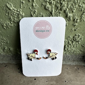 May include: A pair of stud earrings on a white display card featuring the text you can fly design co. Each earring depicts a white goose wearing a red hockey helmet and black skates, holding a brown hockey stick and a black puck in its beak.
