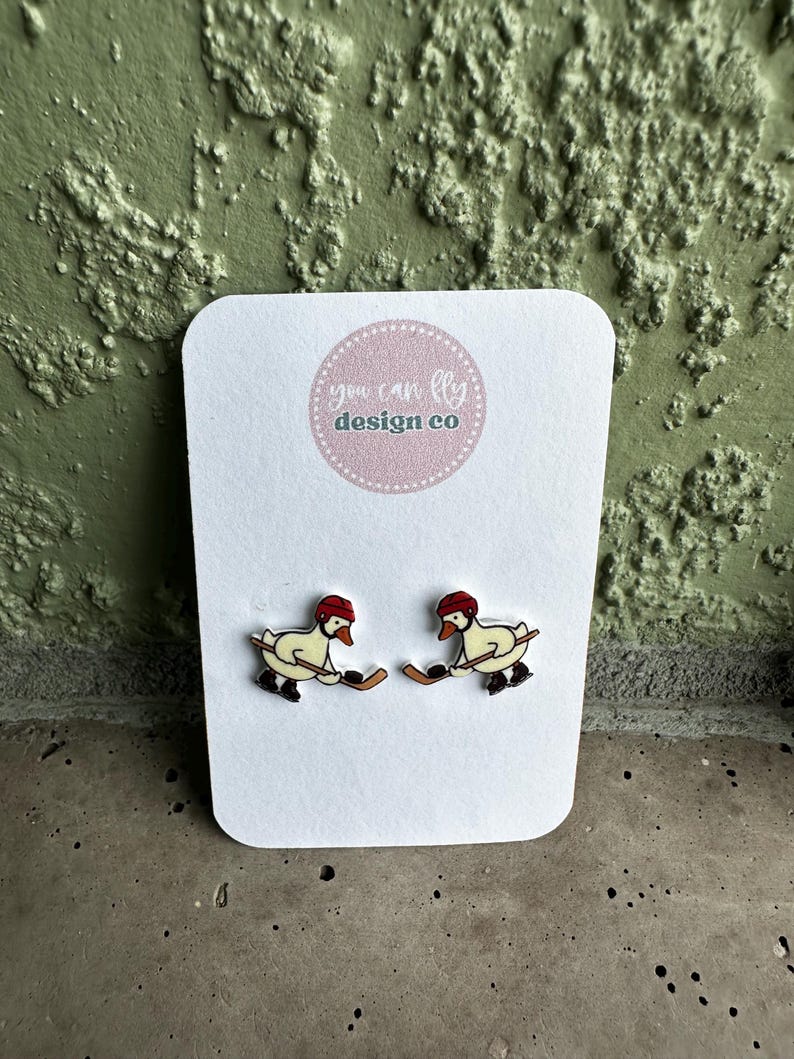 May include: A pair of stud earrings on a white display card featuring the text you can fly design co. Each earring depicts a white goose wearing a red hockey helmet and black skates, holding a brown hockey stick and a black puck in its beak.