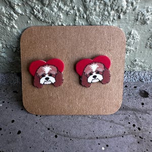 May include: A pair of dog-shaped stud earrings. Each earring features a brown and white dog face with a red heart-shaped background. The earrings are displayed on a brown card.