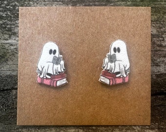 May include: A pair of white ghost earrings with black eyes, each sitting on a stack of three pink books. The books have a white spine and black text.
