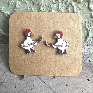 May include: A pair of stud earrings featuring white ducks wearing red hockey helmets and black ice skates, each holding a brown hockey stick with a black puck. The earrings are displayed on a square piece of light brown cardboard with rounded corners.