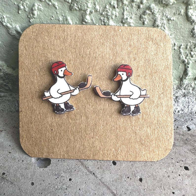May include: A pair of stud earrings featuring white ducks wearing red hockey helmets and black ice skates, each holding a brown hockey stick with a black puck. The earrings are displayed on a square piece of light brown cardboard with rounded corners.