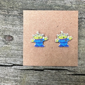 May include: A pair of green alien stud earrings with blue and white details. The earrings are on a brown card.