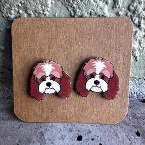 May include: A pair of dog-shaped stud earrings. Each earring features a red and white dog face design with black eyes and a white muzzle. The earrings are displayed on a brown card.