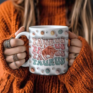 May include: White ceramic mug with a colourful design featuring the word "Taurus" repeated multiple times and a stylised bull graphic. The mug has a floral pattern around the rim.