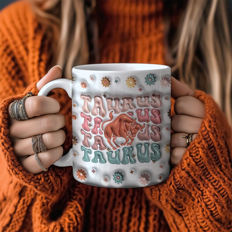 May include: White ceramic mug with a colourful design featuring the word "Taurus" repeated multiple times and a stylised bull graphic. The mug has a floral pattern around the rim.