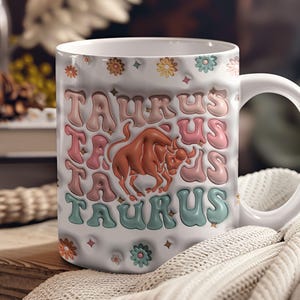 May include: White ceramic mug with a colourful design featuring the word "Taurus" repeated multiple times and a brown bull illustration. The mug has a white handle.