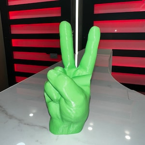 Pode incluir: Uma escultura de mão impressa em 3D de cor verde brilhante fazendo o sinal da paz. A mão está sobre uma superfície branca.