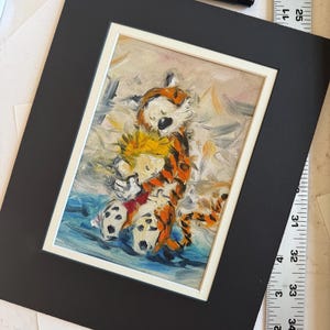 May include: An oil painting of Calvin and Hobbes hugging, set in a white frame on a black mat. The artwork features vibrant colors and brushstrokes, depicting the iconic characters in a tender embrace. A black pen and ruler are in the background.