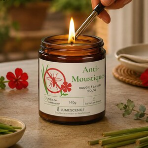 Peut inclure: Une bougie en pot de verre brun avec une étiquette blanche portant l'inscription Anti-Moustiques, Bougie à la cire d'olive, 140g et Lumescence. La bougie présente un motif de moustique dans un cercle rouge et est allumée par un long briquet.