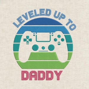Może przedstawiać: Haftowany wzór z kontrolerem do gier wideo wewnątrz niebieskiego, turkusowego i zielonego okręgu. Tekst "LEVELED UP TO DADDY" jest wygięty nad i pod kontrolerem w niebieskiej i różowej nici. Wzór znajduje się na beżowej tkaninie.