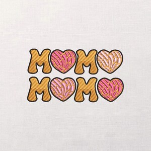 May include: Embroidered design featuring the word "MOM" in gold, outlined in black, with pink and orange heart-shaped accents. The design is on a white fabric background. The letters are in a bold, rounded font.