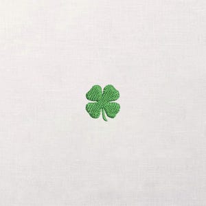 May include: A white fabric with a green embroidered four-leaf clover in the center. The clover is detailed with visible stitching, and the fabric has a subtle texture. The image is a close-up, highlighting the embroidery.