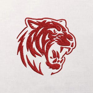 Puede incluir: Una cabeza de tigre bordada en rojo con la boca abierta, mostrando afilados dientes y una expresión feroz. El diseño está cosido sobre una tela blanca, resaltando el detallado trabajo de bordado. La cara del tigre es el punto focal.