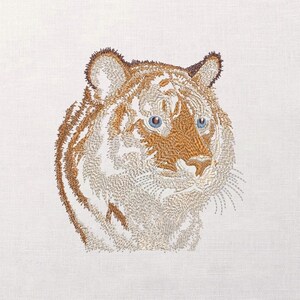 Puede incluir: Cabeza de tigre bordada con rayas marrones y blancas, ojos azules y bigotes detallados. La obra de arte está sobre un fondo blanco, mostrando la intrincada costura y textura del diseño.