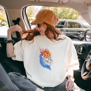 Puede incluir: Sudadera color crema con un gráfico detallado de un león. La melena del león es de color rojo, naranja y azul. La cara del león está intrincadamente diseñada. La sudadera es de estilo casual y cómodo.