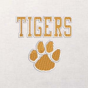 Könnte beinhalten: Gesticktes Design auf weißem Hintergrund mit dem Wort "TIGERS" in goldenen Blockbuchstaben mit weißem Umriss. Darunter ein goldener Pfotenabdruck mit weißem Umriss. Das Design ist wahrscheinlich für Kleidung oder Accessoires.