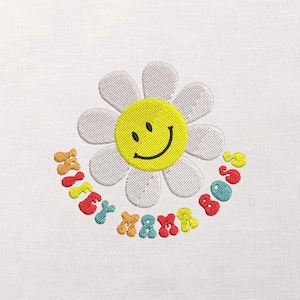 Może przedstawiać: Haftowana stokrotka z żółtym uśmiechniętym środkiem i białymi płatkami. Tekst "Flower Power 80s" jest wyhaftowany w tęczowych kolorach wokół kwiatu, tworząc retro, wesoły design.