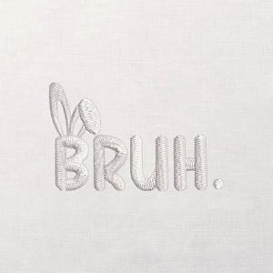 May include: Embroidered text on a white background reads "BRUH." The letter "B" is styled with bunny ears. The embroidery is a light gray color, and the background has a subtle textured appearance.