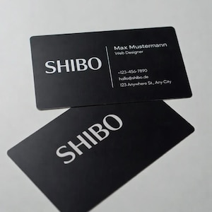 Könnte beinhalten: Zwei schwarze Visitenkarten mit abgerundeten Ecken. Das Wort "SHIBO" ist auf beiden Karten in Weiß gedruckt. Eine Karte zeigt Kontaktinformationen: Max Mustermann, Web Designer, Telefonnummer, E-Mail und Adresse.