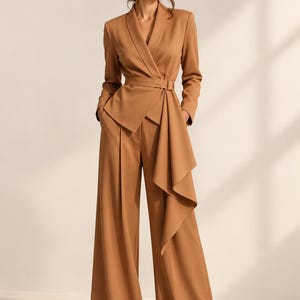 May include: A camel-colored two-piece set consisting of a long-sleeved blazer and wide-leg trousers. The blazer features a shawl collar, a wrap-style front with a matching fabric belt, and a cascading side panel. The trousers have a high waist.