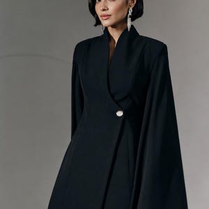 May include: A black blazer dress featuring a high collar, a single decorative crystal button at the waist, and integrated cape-style sleeves that drape down the sides. The garment has a structured silhouette and a smooth, solid black fabric finish.