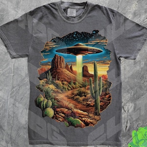 May include: A dark gray t-shirt with a graphic print of a flying saucer over a desert landscape. The graphic features a desert scene with cacti, mountains, and a starry sky.