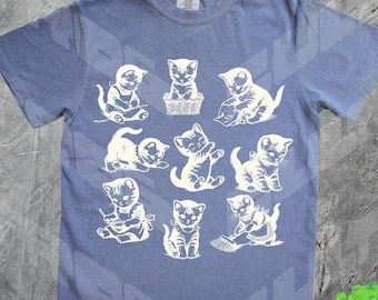 Könnte beinhalten: Ein hellblaues T-Shirt mit einem weißen Grafikdruck von 10 Kätzchen in verschiedenen Posen. Die Kätzchen tragen alle verschiedene Outfits und Accessoires.