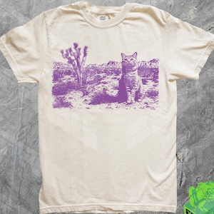 May include: A white t-shirt with a purple print of a cat sitting in a desert landscape. The cat is looking to the left. The print includes a Joshua tree in the background.