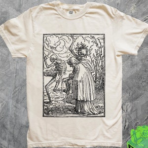 May include: A white t-shirt with a black and white woodcut print of Death and a woman. The print depicts Death as a skeleton holding a scythe and the woman is wearing a long robe. The print is framed by a black border.
