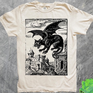 May include: Cream-coloured t-shirt with a black graphic of a winged cat on a castle. The cat has bat-like wings and is set against a detailed castle backdrop. The shirt has a crew neck and short sleeves.