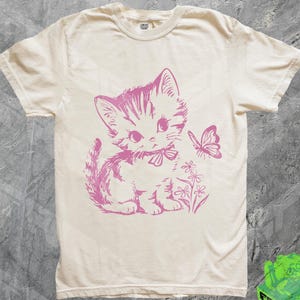 May include: Cream-colored t-shirt featuring a pink illustration of a kitten with a bow, a butterfly, and flowers. The shirt has short sleeves and a crew neck. The text "CONVERT FASHION" is printed on the collar.
