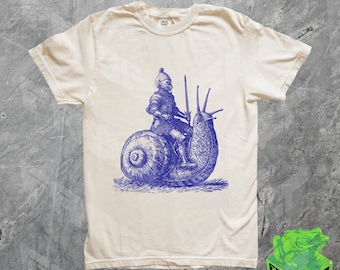 May include: A white t-shirt with a blue illustration of a knight riding a snail. The knight is wearing armor and holding a sword. The snail is large and has a detailed shell.